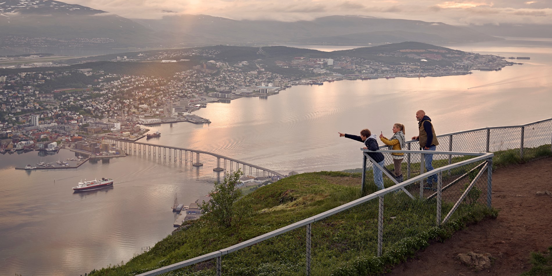 Three children stand behind the fence on top of the mountain in the city of Tromsø. They are overlooking the city of Tromsø.