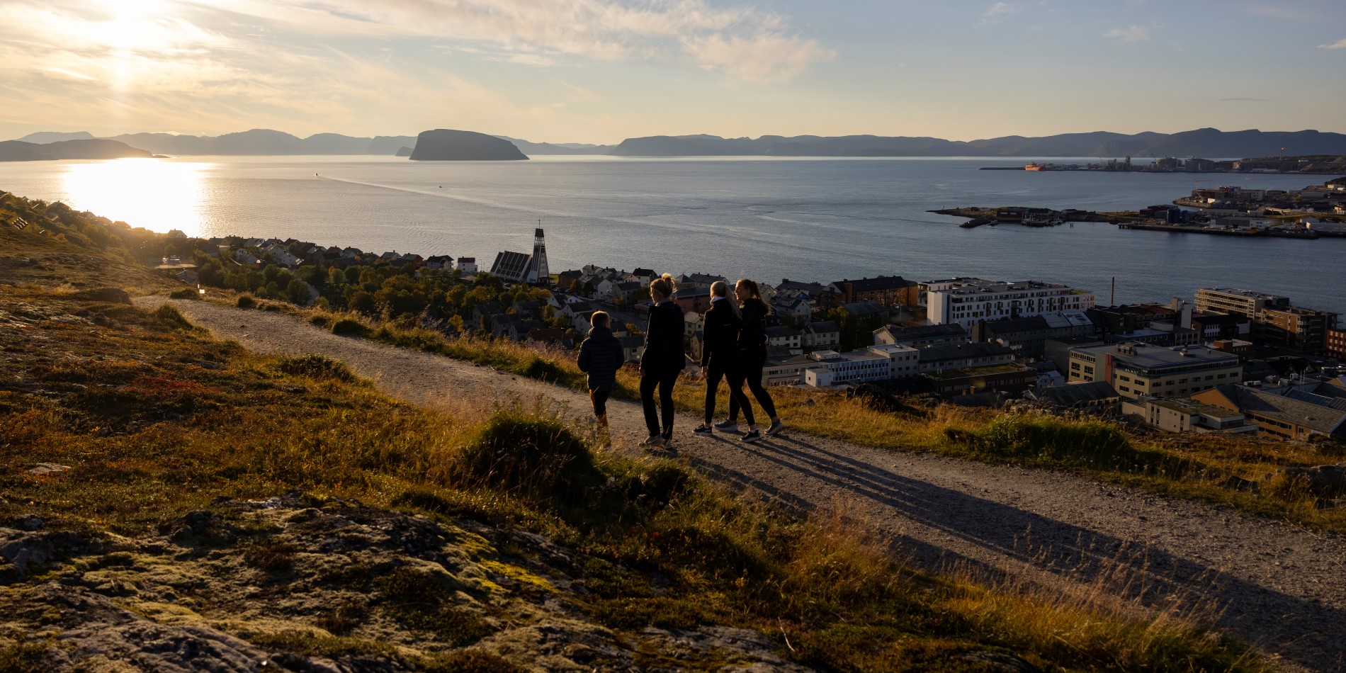 It’s sunset and four people are walking on the hillside. Below them, they can see the town of Hammerfest, Norway. 